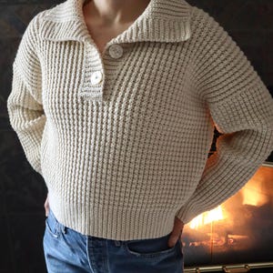 May include: A cream-colored waffle knit sweater with a button-up collar and long sleeves. The sweater is cropped and has a relaxed fit.