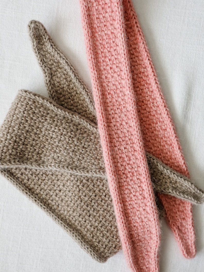 K&ouml;nnte beinhalten: Zwei geh&auml;kelte Stirnb&auml;nder, eines braun und eines rosa, liegen auf einer wei&szlig;en Oberfl&auml;che. Die Stirnb&auml;nder sind mit einer einfachen Masche gearbeitet und haben eine weiche, flauschige Textur.