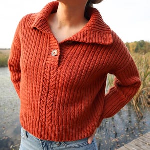 May include: An orange-red knitted sweater with a collared neckline and a single wooden button closure. The sweater features a cable knit design down the center and ribbed detailing. The model is wearing blue jeans.