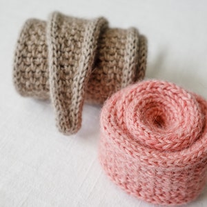 K&ouml;nnte beinhalten: Zwei Kn&auml;uel Wolle, eines beige und eines rosa, sind aufgerollt und auf einer wei&szlig;en Oberfl&auml;che platziert.