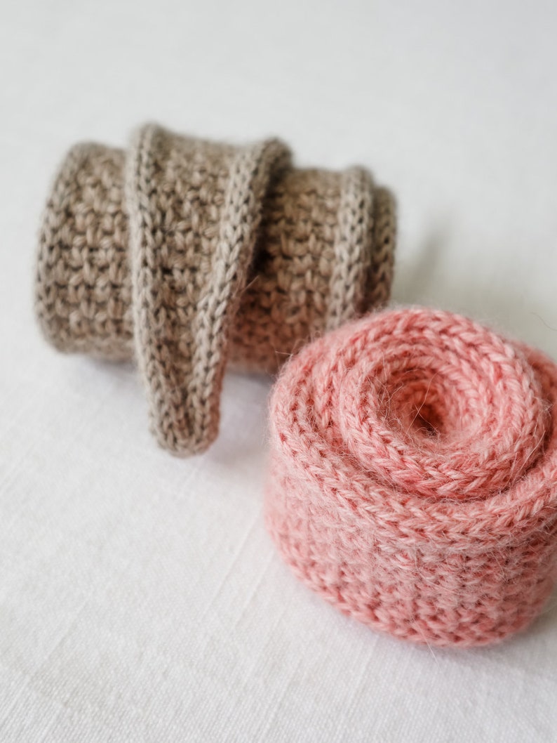 K&ouml;nnte beinhalten: Zwei Kn&auml;uel Wolle, eines beige und eines rosa, sind aufgerollt und auf einer wei&szlig;en Oberfl&auml;che platziert.