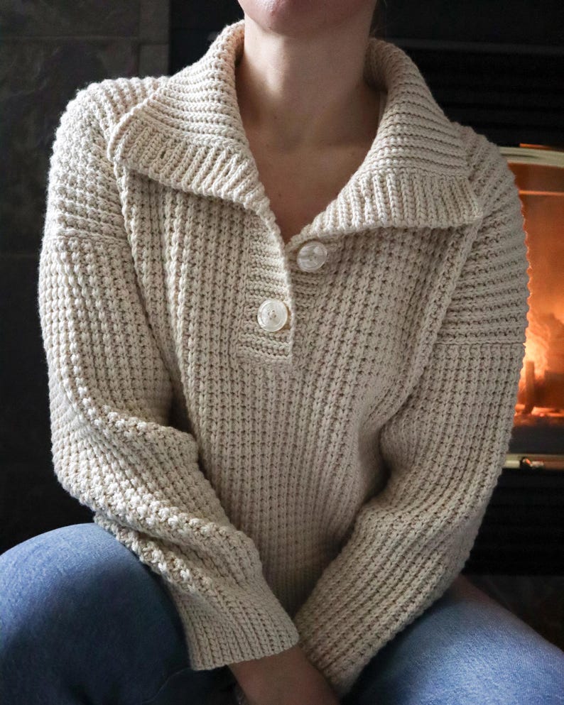 May include: A cream-colored knitted sweater with a button-up collar and long sleeves. The sweater is made of a textured yarn and has a relaxed fit.
