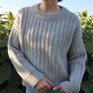 May include: A light grey, chunky knit jumper with a ribbed crew neck and sleeves. The jumper has a textured, vertical pattern and is worn with blue jeans. The background features sunflowers and a clear sky.