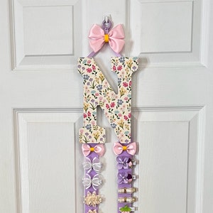 May include: A decorative hair bow holder shaped like the letter "N". The top of the "N" is floral patterned. Two lilac ribbons hang down, each with decorative bows and clips holding various hair accessories. A pink bow is at the top.