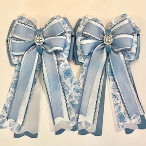 May include: Two light blue hair bows with white floral patterns and silver accents. Each bow features a central decorative emblem and long, flowing ribbons. The bows are set against a white background, with white roses in the background.