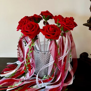May include: A bouquet of red rose wands with white and burgundy ribbon streamers and green leaves. The wands are in a clear glass vase.