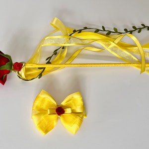 May include: A yellow and red wand with a red rose and green leaves on the top. The wand has yellow ribbons attached to it. There is also a yellow bow with a red rose in the center.