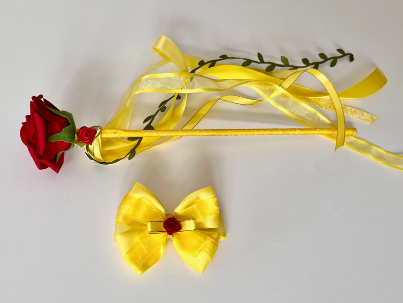 May include: A yellow and red wand with a red rose and green leaves on the top. The wand has yellow ribbons attached to it. There is also a yellow bow with a red rose in the center.
