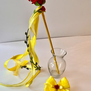 May include: A yellow ribbon wand with a red rose on top. The wand is leaning against a clear glass vase. There is also a yellow bow with a red rose in the center.