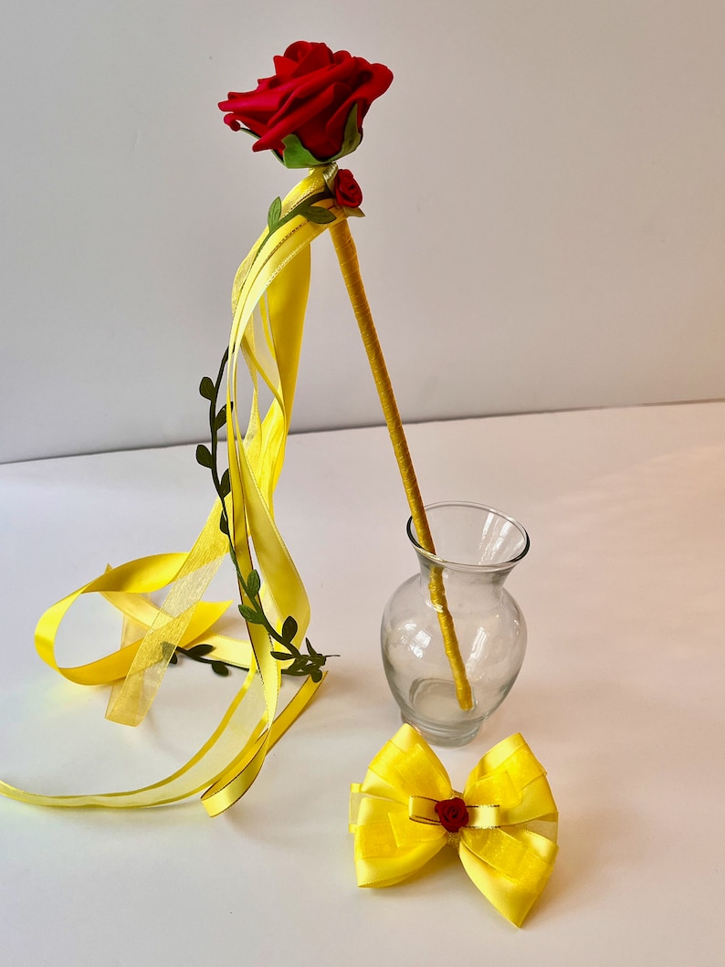 May include: A yellow ribbon wand with a red rose on top. The wand is leaning against a clear glass vase. There is also a yellow bow with a red rose in the center.