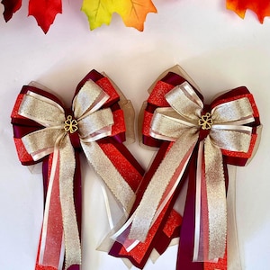 May include: Two decorative bows with burgundy, gold, and red ribbons. Each bow features a gold-colored floral embellishment at the center. The bows have long, layered streamers in coordinating colors. Autumn leaves are in the background.