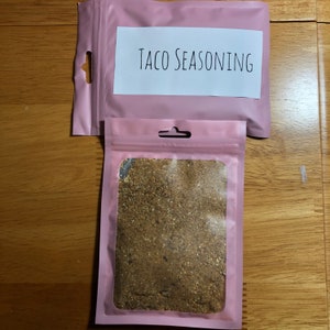Pode incluir: Um saco rosa lacrável com uma janela transparente que mostra tempero para tacos castanho no interior. O saco tem um rótulo que diz "Taco Seasoning".