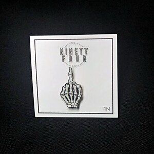 May include: A white skeleton hand giving the middle finger, on a white card with black text that reads "The Ninety Four Shop" and "Pin".