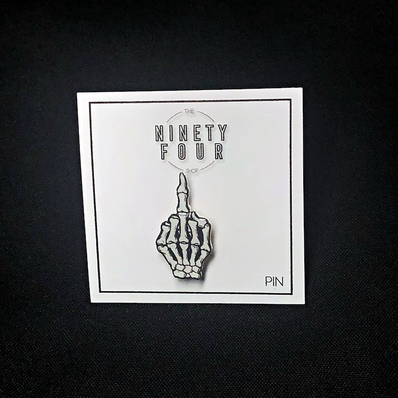 May include: A white skeleton hand giving the middle finger, on a white card with black text that reads "The Ninety Four Shop" and "Pin".
