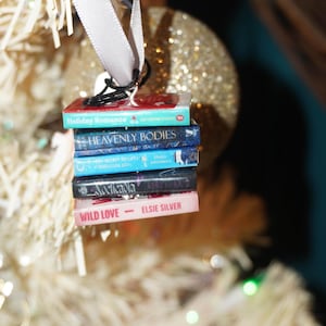 May include: A stack of miniature book ornaments with titles like "Holiday Romance" and "Wild Love" hangs from a white ribbon. The books are in various colors, including red, blue, and pink, and are suspended in front of a blurred Christmas tree.