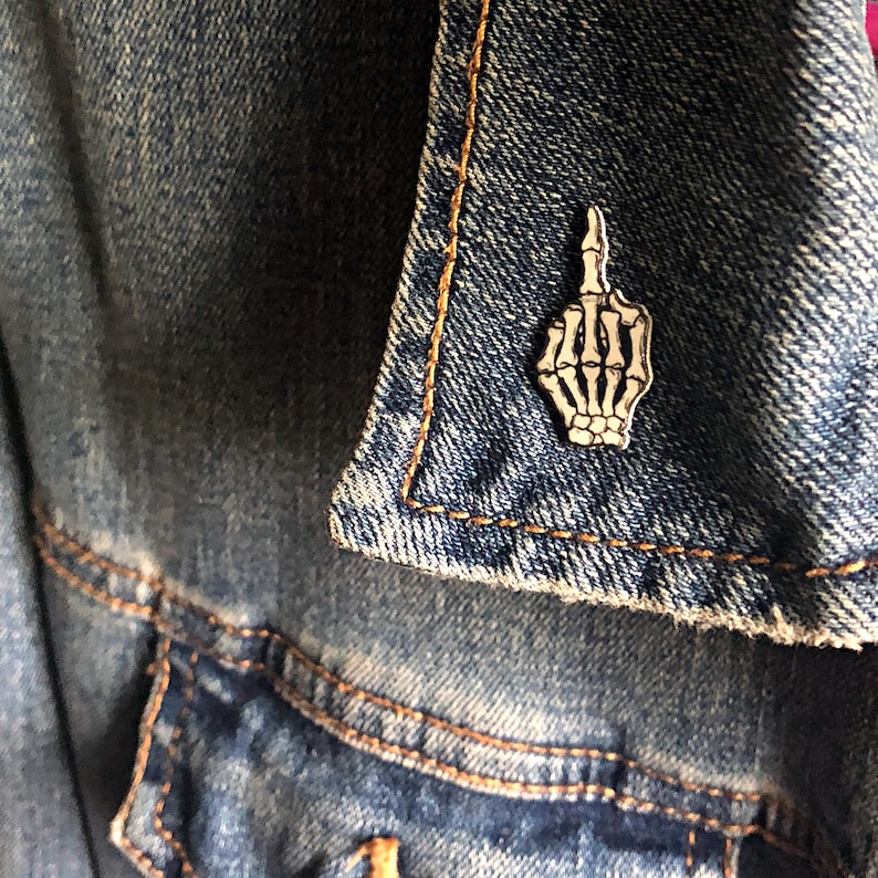 May include: A white skeleton hand giving the middle finger, depicted as a metal pin on a blue denim jacket.