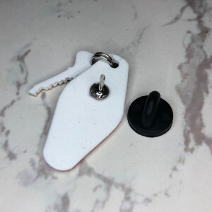 May include: White enamel pin shaped like a hotel key with a black rubber clutch back.