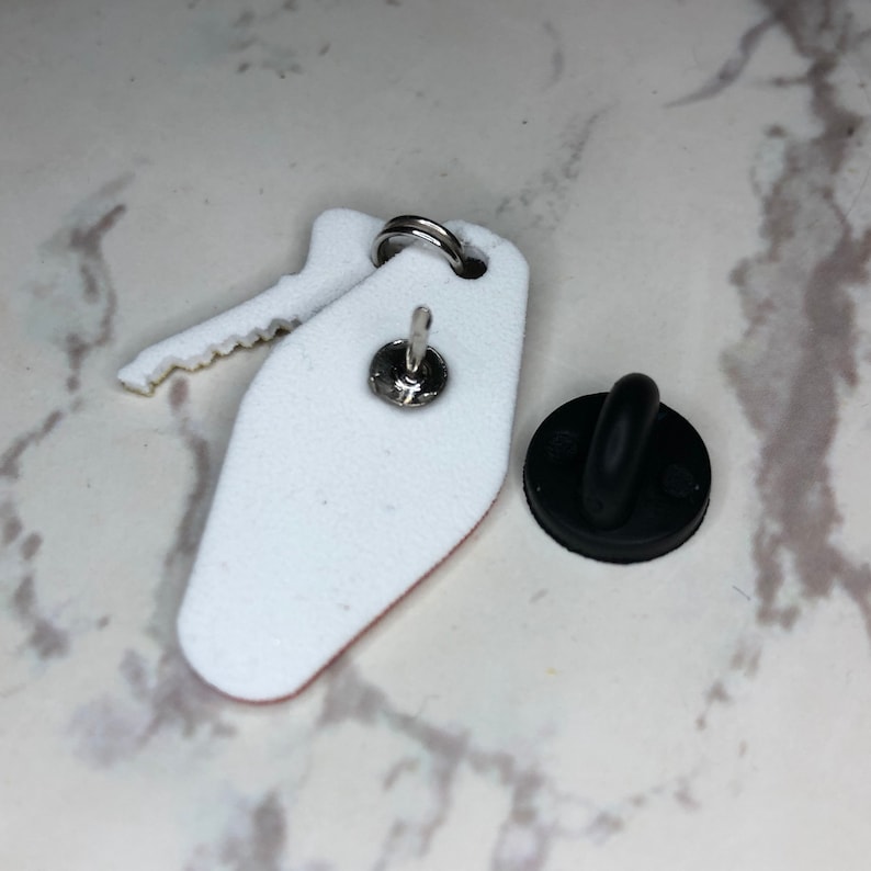 May include: White enamel pin shaped like a hotel key with a black rubber clutch back.