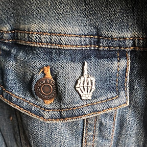 May include: A silver skeleton hand pin with a middle finger raised, pinned to a blue denim jacket with a brown button that says "Old Navy - San Francisco, CA."