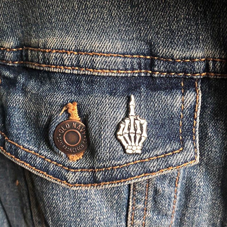 May include: A silver skeleton hand pin with a middle finger raised, pinned to a blue denim jacket with a brown button that says "Old Navy - San Francisco, CA."