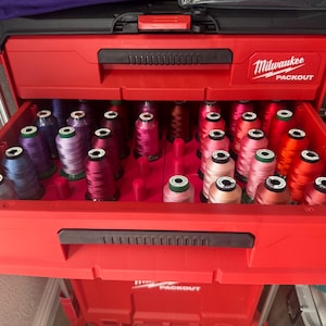 May include: A red Milwaukee Packout storage box with a drawer filled with colorful spools of thread. The thread colors include shades of blue, purple, pink, orange, and red. The box is open, revealing the organized thread spools.