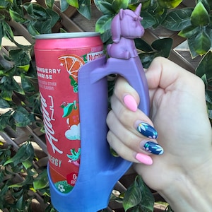May include: A purple 3D-printed beverage can holder featuring a small unicorn figure on the top of the handle. The holder is designed to fit a standard slim aluminum can with a red and orange label that reads Strawberry Sunrise and Alani Nu Energy.
