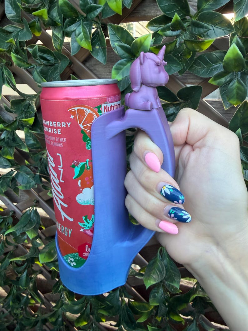May include: A purple 3D-printed beverage can holder featuring a small unicorn figure on the top of the handle. The holder is designed to fit a standard slim aluminum can with a red and orange label that reads Strawberry Sunrise and Alani Nu Energy.