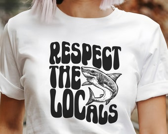 May include: White t-shirt with a black graphic that says "Respect the Locals" with a shark illustration.