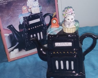 May include: Black ceramic teapot shaped like an old-time radio with a white cat figurine on top. The teapot has a black and white dial with the words "On The Air" and a clock on the side. The teapot is 6 oz capacity and made in China.