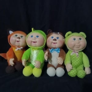 May include: Four Cabbage Patch Kids dolls dressed in animal costumes. The dolls have blue eyes and rosy cheeks. Costumes include a fox, a lizard, a bear, and a frog. The dolls are plush and have a soft appearance.