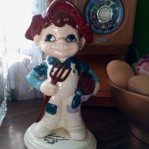 May include: A ceramic figurine of a boy wearing a red hat and blue overalls holding a pitchfork. The figurine is standing on a white base with the inscription "The Woodland Wildlife Foundation Class of '85 1985".