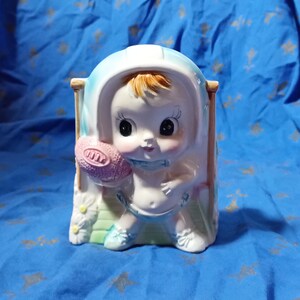 May include: A ceramic planter shaped like a baby holding a pink ball. The baby is wearing a blue hooded onesie and has large eyes. The planter has a white background with green and blue accents.