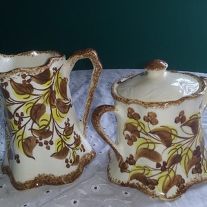 May include: Cream-colored ceramic pitcher and lidded sugar bowl set. Both pieces feature a brown and yellow floral design with a scalloped rim. The pitcher has a curved handle, and the sugar bowl has a matching lid with a small knob.