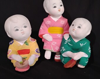 May include: Three ceramic dolls wearing traditional Japanese clothing. The doll on the left is wearing a yellow kimono with red and green accents. The doll in the middle is wearing a pink kimono with a yellow sash. The doll on the right is wearing a green kimono with white accents.