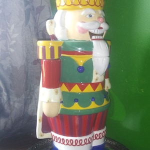 May include: A ceramic nutcracker figurine with a colorful, multi-layered design. The nutcracker is wearing a purple and yellow crown, a red and green jacket, and blue boots. The nutcracker is standing on a silver tray with a mirrored surface.