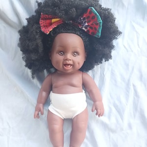 May include: A black doll with brown eyes and a big smile, wearing a white diaper and a colorful bow in her afro hair.