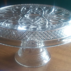 May include: A clear glass cake stand with a decorative, raised, diamond pattern around the edge of the top plate and base. The top plate has a raised, floral design.