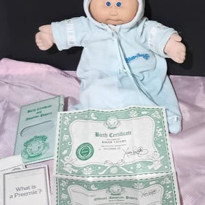 May include: A Cabbage Patch Kids doll wearing a light blue hooded outfit. The doll has a soft, fabric body and a painted face with blue eyes. Included are birth and adoption papers, and a booklet titled "What is a Preemie?"