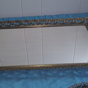 May include: A gold-colored rectangular tray with a mirrored surface and an ornate, decorative frame. The tray has four small feet.