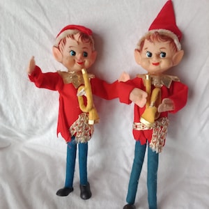 May include: Two vintage elf figurines. Each elf wears a red jacket, hat, and blue pants. They have gold-colored collars and belts. Each elf holds a yellow trumpet. The elves have rosy cheeks and are smiling.