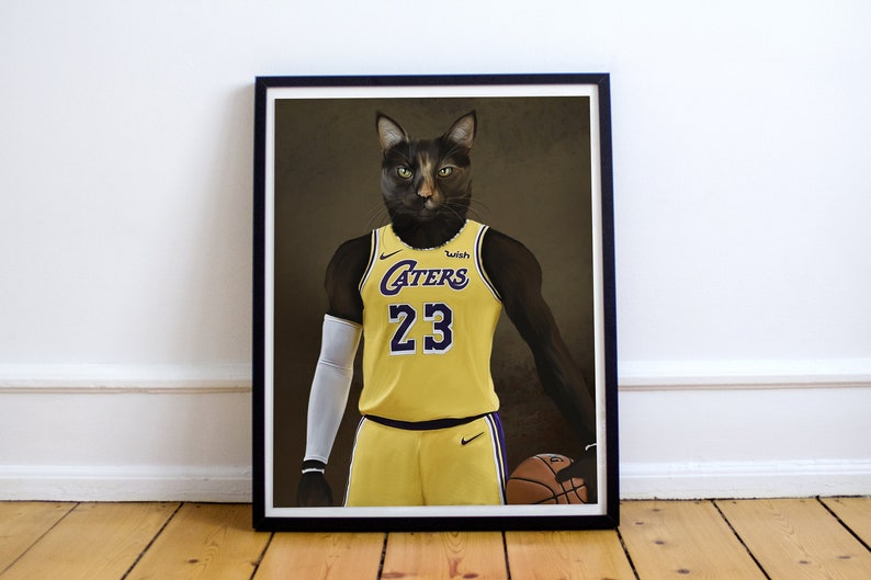 May include: Framed print featuring a cat wearing a yellow basketball jersey with the number 23. The jersey has purple accents and the word "Caters". The cat is holding a basketball. The print is in a black frame.