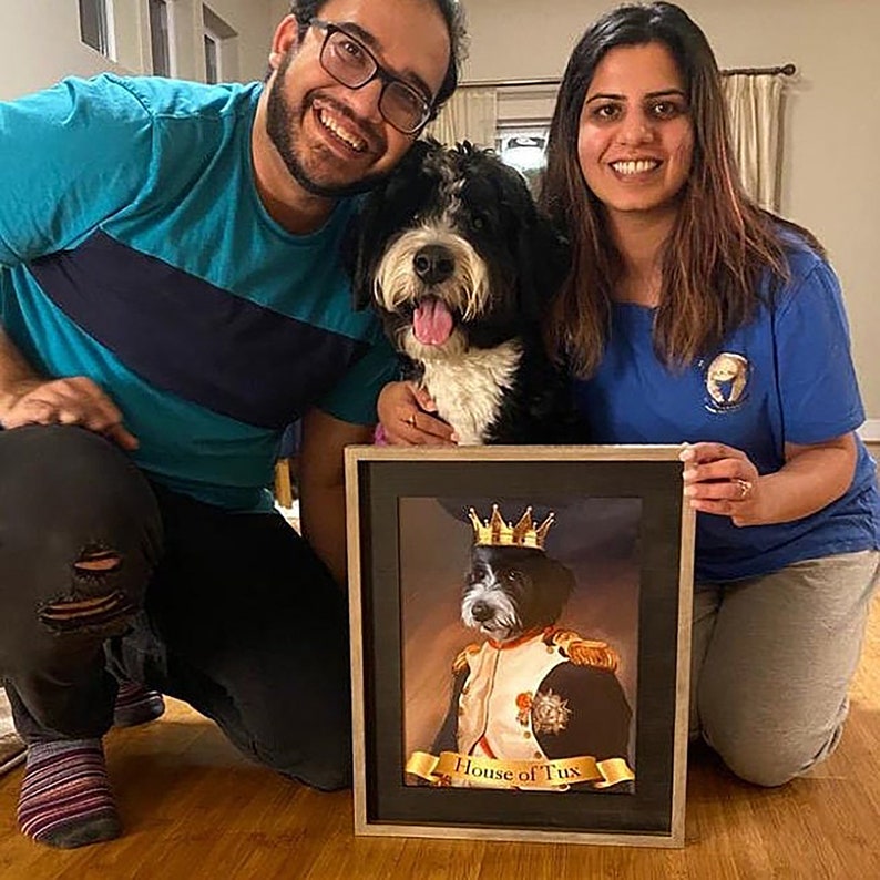 May include: A framed portrait of a black and white dog wearing a crown and military-style jacket, with the text "House of Tux". The portrait is held by a couple, with the dog in between them.