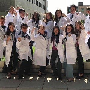 May include: A group of people wearing white lab coats and dark pants, posing with flexed arms. Each person has a patterned scarf. The background includes a building and green grass.