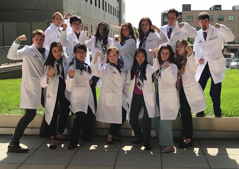 May include: A group of people wearing white lab coats and dark pants, posing with flexed arms. Each person has a patterned scarf. The background includes a building and green grass.