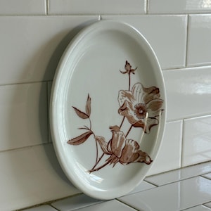 May include: Oval white ceramic plate with a brown floral design. The plate features a detailed illustration of a flower with petals and leaves. The plate is a vintage style and is suitable for serving food.