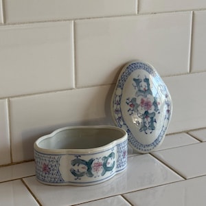 May include: A small, open-top ceramic container and a matching lid. The pieces are white with a blue floral and geometric pattern. The container has a curved shape with a scalloped edge. The lid is oval-shaped.