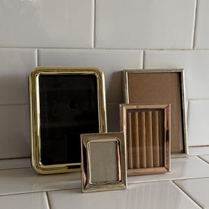 May include: Four decorative picture frames of varying sizes and styles. The frames include gold, silver, and rose gold finishes. The frames are empty, ready to display photos or artwork. The frames are arranged on a white tiled surface.