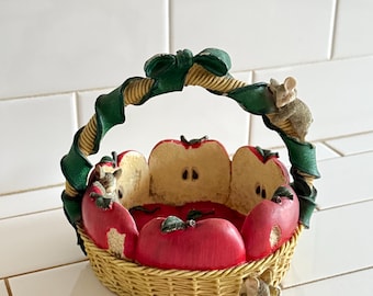 Puede incluir: Una cesta decorativa con una base de textura tejida y un asa envuelta en una cinta verde. El exterior está adornado con mitades de manzanas rojas y pequeños ratones esculpidos. El interior muestra la pulpa clara de las manzanas cortadas.