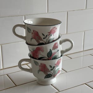 May include: Three stacked white teacups with a floral design. Each cup features a pink rose pattern with green leaves and a dark blue rim. The cups have a handle and a small pedestal base.