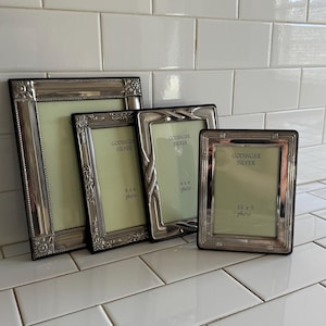 May include: A set of four rectangular silver-toned picture frames with varying decorative borders. Each frame contains a light green insert with the text Godinger Silver and the photo size, including 4x6 inches and 3.5x5 inches, printed on the front.